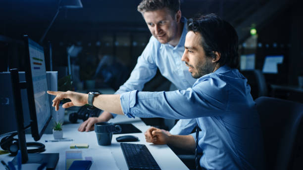 Ambitious and Confident Businessman Uses Desktop Computer, His Project Manager Explains Specific Tasks, Account Handling and Strategic Moves. Professional People Late at Night in Big Corporate Office